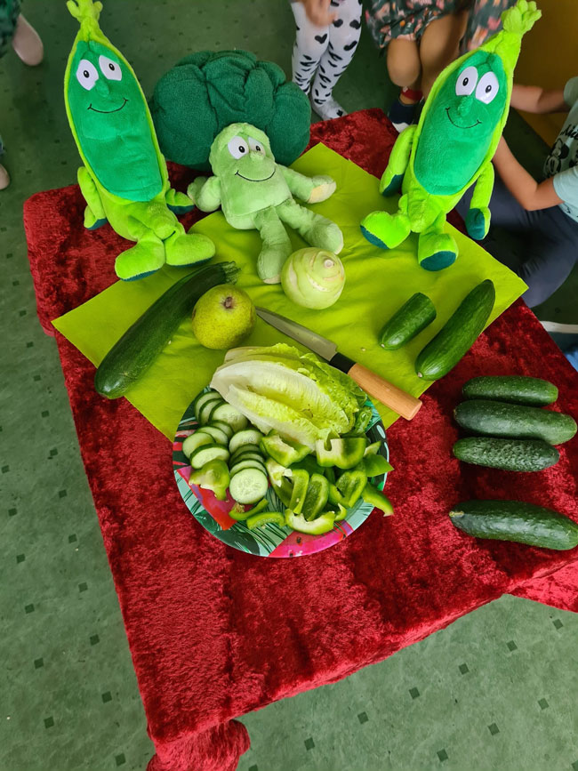 Bordowy materiał, zielona papryka, ogórki, kalarepka, gruszki, talerz z pokrojonymi ogórkami, zielone maskotki: dwa ogórki.