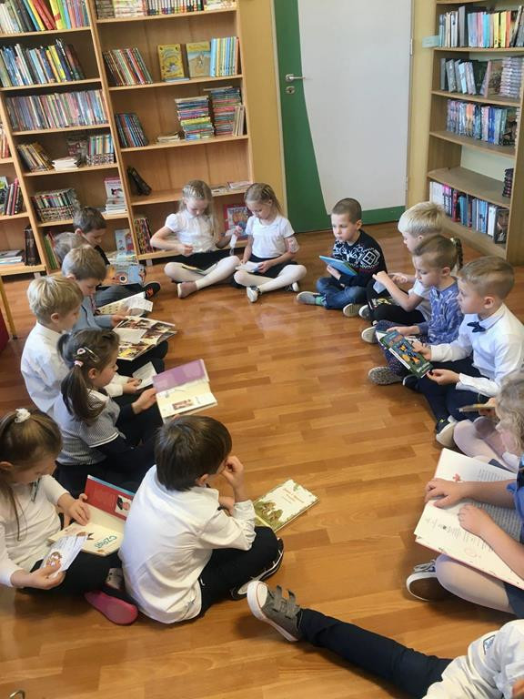 Uczniowie w bibliotece. Siedzą na podłodze, w dłoniach trzymają książki. Z tyłu stoją regały z książkami.