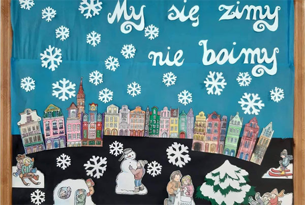 Tablica - niebieskie tło, biały napis 'My się zimy nie boimy', białe śnieżynki, kolorowe domy, bałwan, choinka obsypana śniegiem.