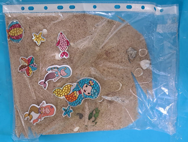 Praca dziecka - w woreczku strunowym piasek z plaży, muszelki, naklejone kolorowa rybka, rozgwiazdy, 3 syrenki.