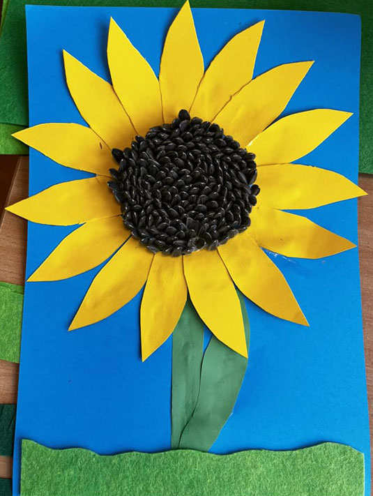 Niebieskie tło, żółty kwiat słonecznika wykonany z papieru, środek - pestki słonecznika.