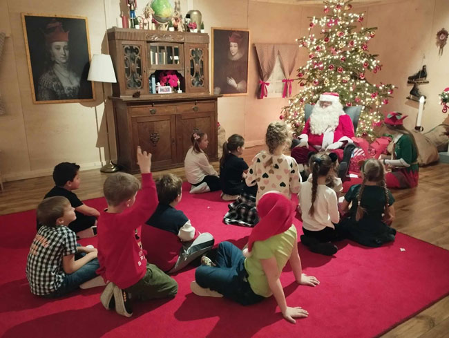 Dzieci siedzące na czerwonym dywanie, Mikołaj, choinka z ozdobami.
